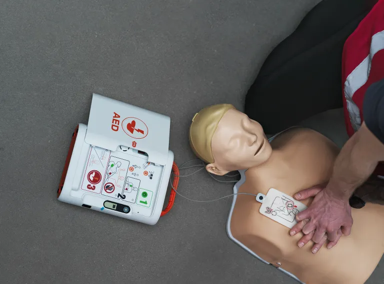 Hænder giver hjertemassage på en øvelsesdukke ved siden af en hjertestarter